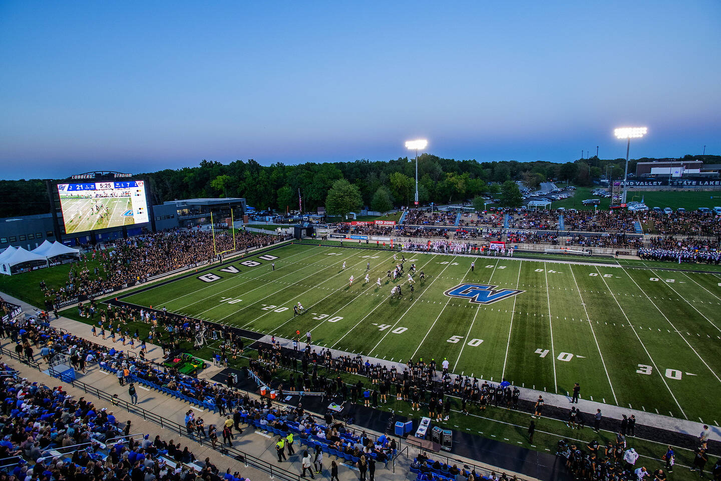 An aerial view of a football game at dusk, with players on the field, a large crowd in the stands, and a scoreboard displaying the score.
